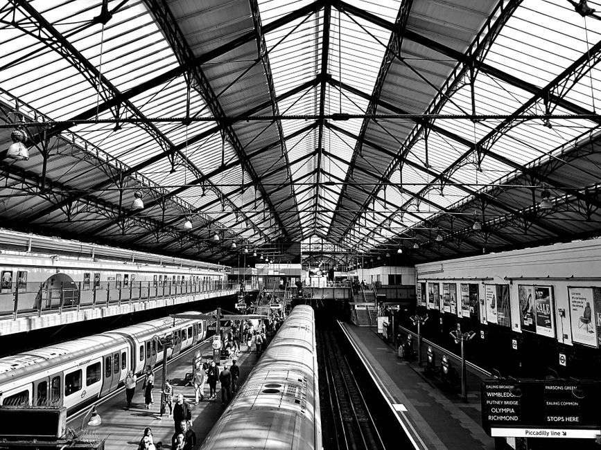A black and white photograph of Bow Road station showing the station's high, arched metal and glass roof structure, which allows natural light to illuminate the platform area. The scene includes two train tracks with trains on either side, busy with passengers walking along the platform, some waiting or boarding. On the right side, a wall is decorated with several posters and signs, while underneath the roof, overhead lights are visible. The clean, maintained surfaces and organized layout exemplify efficient metro station design, with a focus on commuter safety and comfort, highlighting the importance of surface cleaning and hygiene in transportation hubs. This setting is typical of the professional cleaning measures likely managed by Bow Cleaners, offering expert hygiene and surface cleaning services for public and commercial spaces.