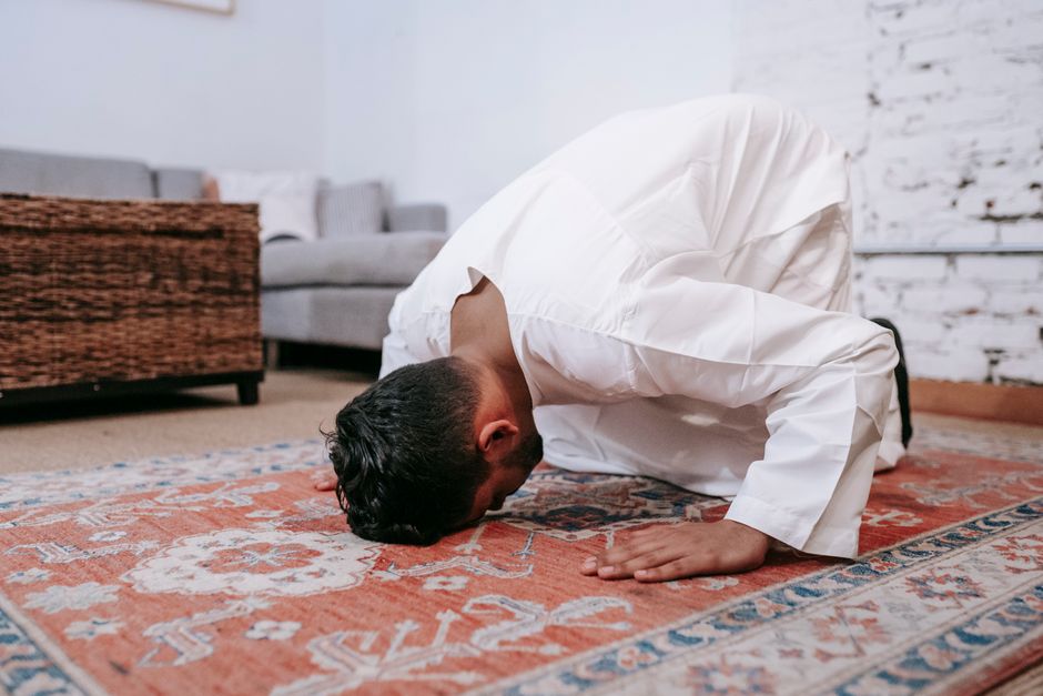 A man dressed in a white shirt and dark trousers is performing an act of Islamic prayer (Sujud) on a patterned, traditional rug with intricate designs in orange and blue hues. The room features natural lighting, a light-colored brick wall, and visible furnishings including a woven basket and a light grey sofa in the background. The polished floors are clean and reflect the light, indicating good surface maintenance. The setting appears to be a domestic space, emphasizing the importance of regular cleaning and upkeep to maintain hygiene and a peaceful environment. This image aligns with the theme of surface cleaning and hygiene, as seen on the Bow Road station quick carpet cleaning options nearby page of bowcleaners.org.uk.