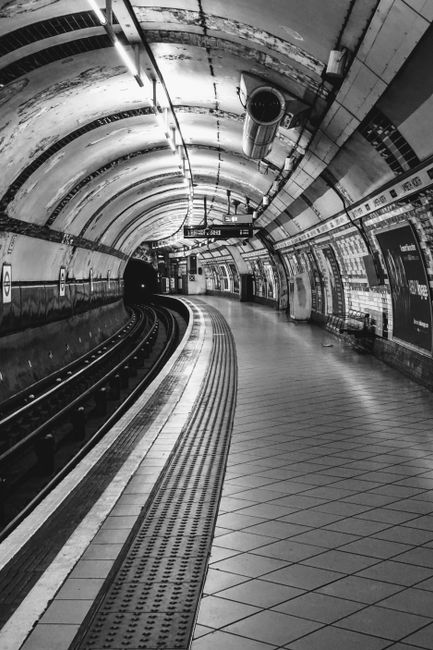 A black and white photograph of Bow Road station showing the station's high, arched metal and glass roof structure, which allows natural light to illuminate the platform area. The scene includes two train tracks with trains on either side, busy with passengers walking along the platform, some waiting or boarding. On the right side, a wall is decorated with several posters and signs, while underneath the roof, overhead lights are visible. The clean, maintained surfaces and organized layout exemplify efficient metro station design, with a focus on commuter safety and comfort, highlighting the importance of surface cleaning and hygiene in transportation hubs. This setting is typical of the professional cleaning measures likely managed by Bow Cleaners, offering expert hygiene and surface cleaning services for public and commercial spaces.