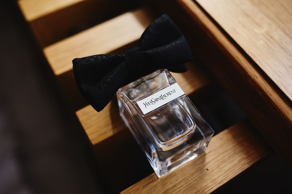 A clear glass Yves Saint Laurent perfume bottle with a black bow tied around its neck, placed on a wooden surface with visible grain, adjacent to a wooden structure. The bottle's surfaces are clean and shiny, reflecting light, indicating a well-maintained condition. The surrounding area features warm lighting that highlights the smooth, polished finish of the wood, suggesting careful surface cleaning and attention to detail, consistent with professional domestic cleaning practices promoted on bowcleaners.org.uk for end of tenancy hygiene and sanitisation.