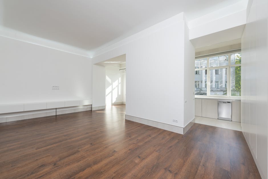 A spacious, well-lit room with white painted walls and large windows that allow natural light to illuminate the space. The flooring consists of polished, dark wood laminate, appearing clean and free of dust or debris. Near the window, there is a small white built-in cabinet set into the wall. The room features a minimalistic design with smooth surfaces and tidy edges, indicative of recent deep cleaning or preparation for final tenant handover. The lighting emphasizes the pristine condition of the surfaces, and the overall atmosphere suggests a professional cleaning service has been applied by Bow Cleaners, aligning with the topic of end of tenancy cleaning tips for Bow E3.