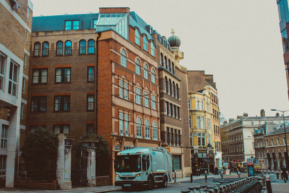 A street scene featuring a mix of historic and modern buildings, including a prominent red brick structure with numerous multi-pane windows, adjacent to lighter-colored and yellow façades with decorative architectural details, under a pale sky. The street is lined with parked bicycles, bollards, and a white waste collection truck driving past. The area appears clean and well-maintained, with clear pavements and minimal visible debris. The image captures the urban environment near Roman Road Market, showcasing diverse architectural styles and the importance of surface cleaning and maintenance in residential and commercial settings, as promoted by bowcleaners.org.uk for domestic cleaning and deep cleaning services.