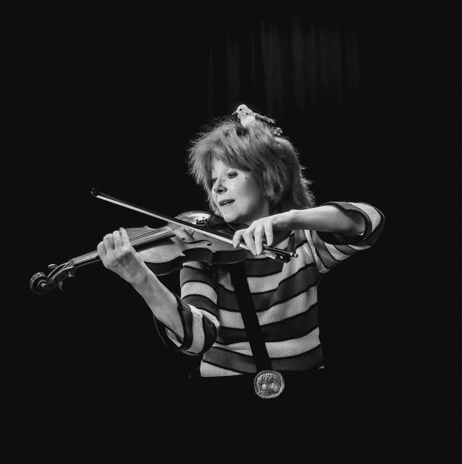 The image shows a woman with tousled, shoulder-length hair playing a violin with focused expression, standing against a dark background. She is wearing a striped long-sleeve shirt and a large medal around her neck. The lighting highlights her face and the violin, emphasizing the performance setting. In the context of Victoria Park event cleanup services for Bow party venues, this image suggests the importance of maintaining a clean and hygienic environment for public gatherings, with professional cleaning services provided by bowcleaners.org.uk to ensure surfaces such as floors, counters, and other areas are thoroughly cleaned and sanitised for safe and enjoyable events.